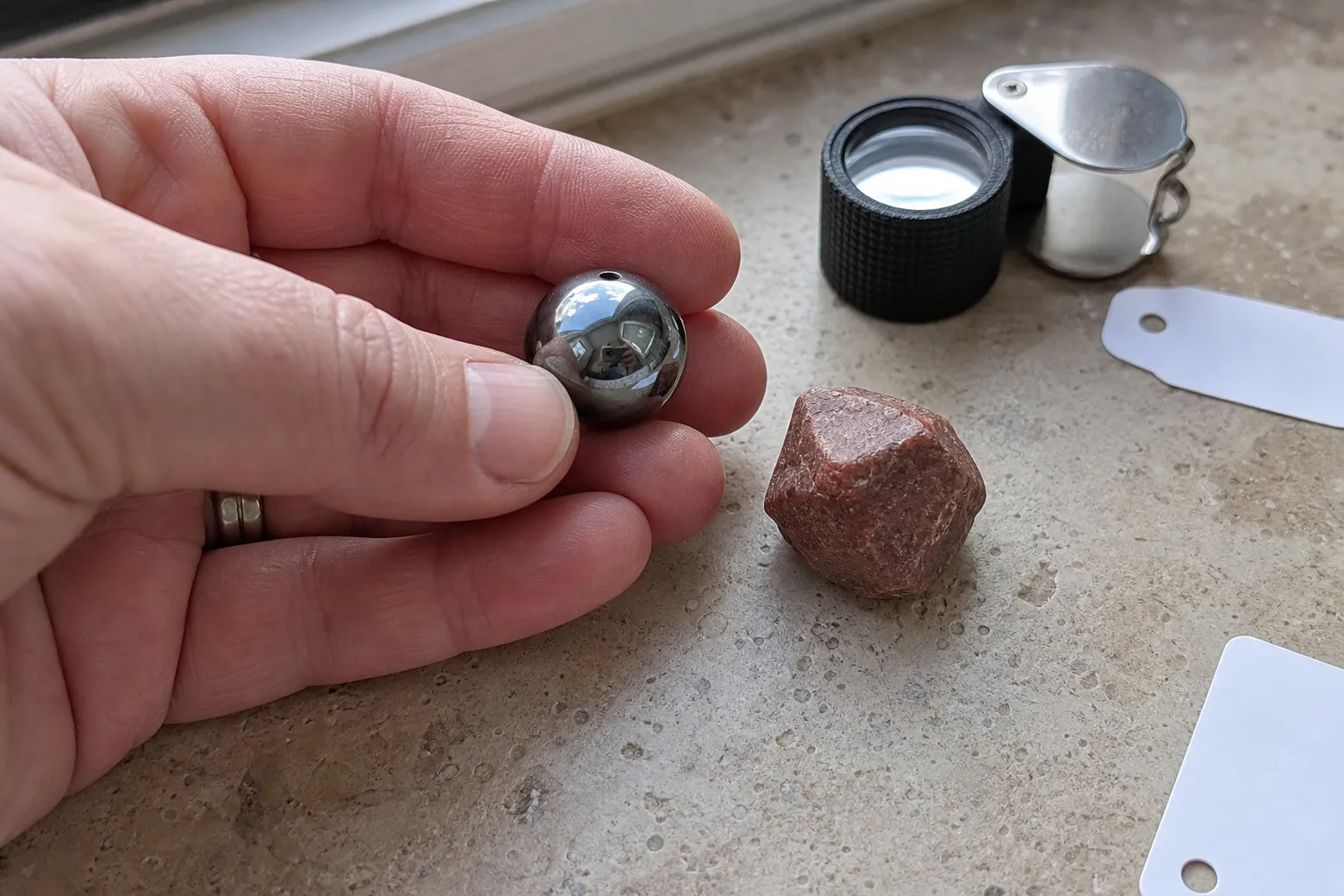 A hand comparing a metallic gray hematite bead and a reddish mineral piece by size and weight impression